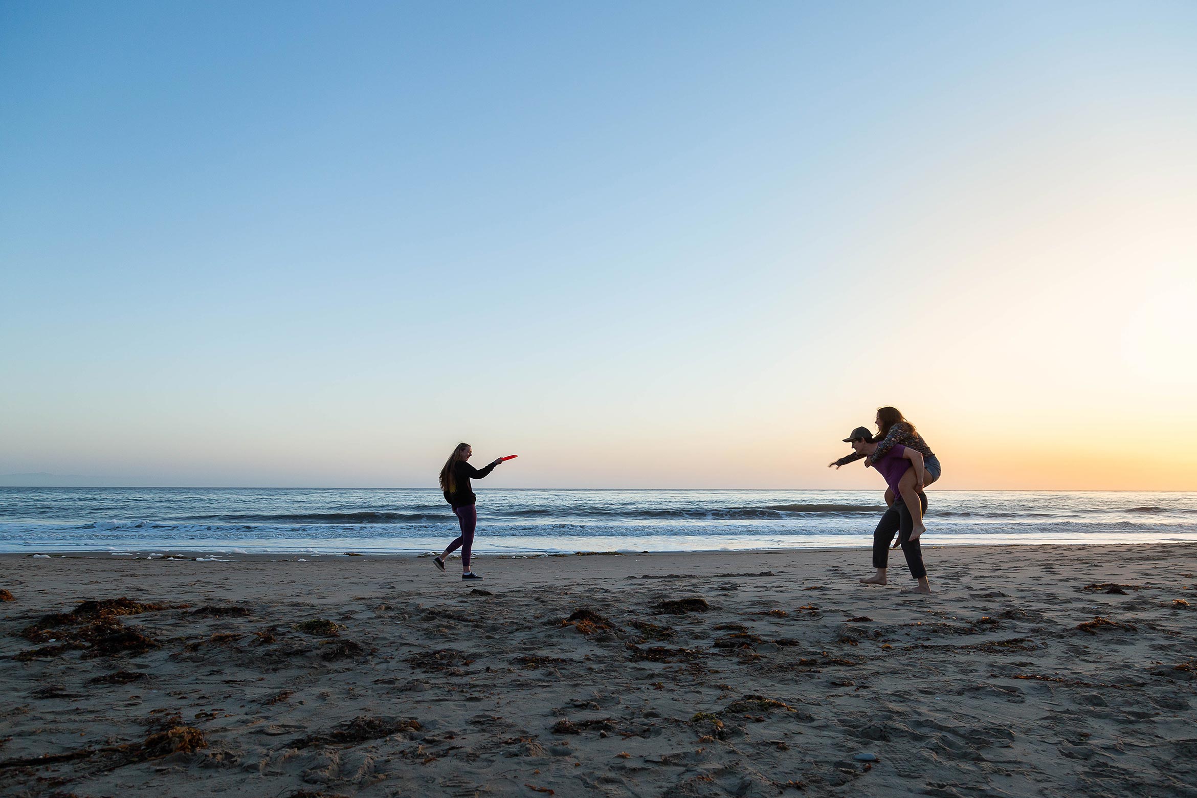 Three students play frisbee on the beach at dusk.