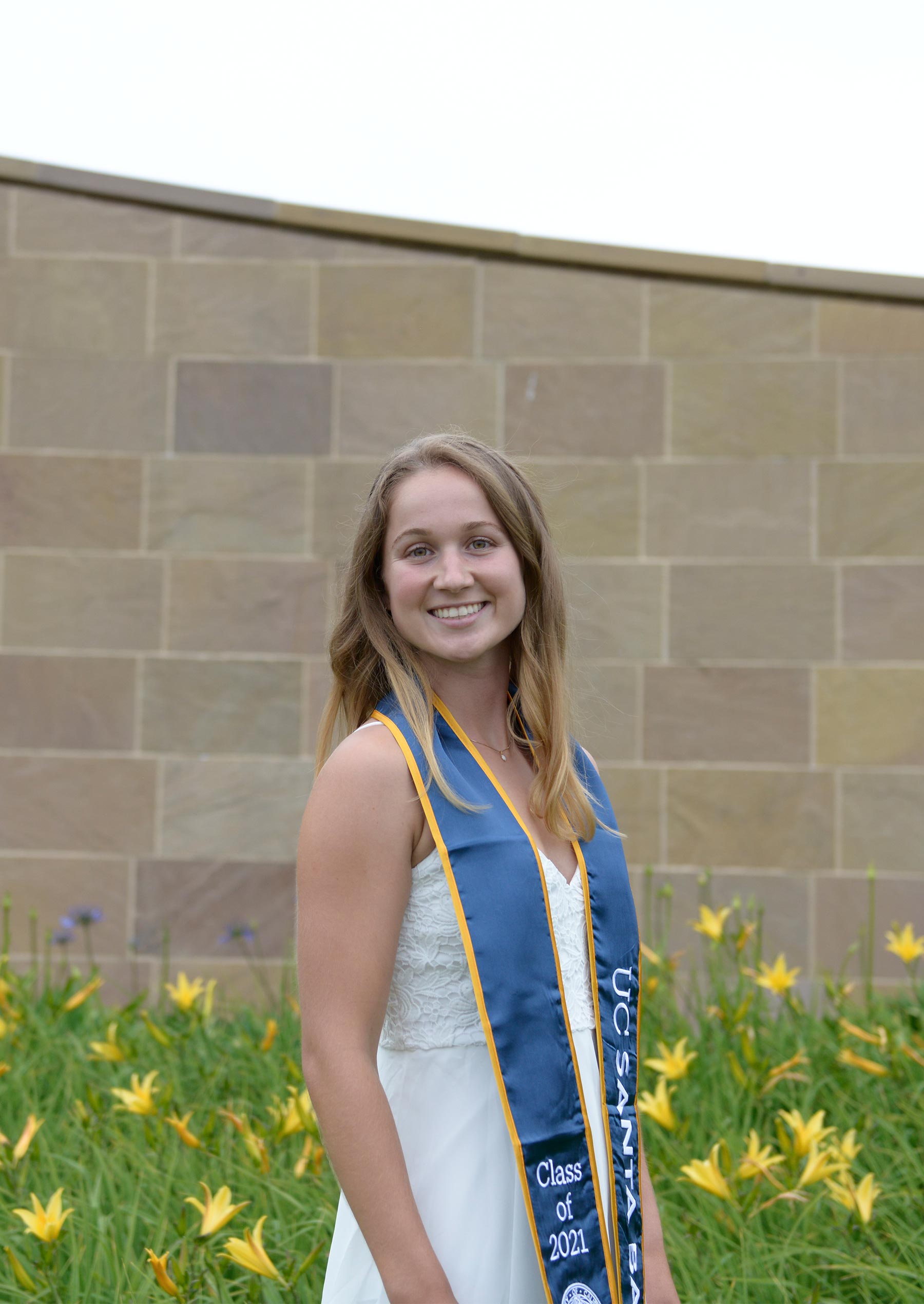Grace Wiley wears a Class of 2021 stole and stands among the flowers in front of Henley Gate at UC Santa Barbara
