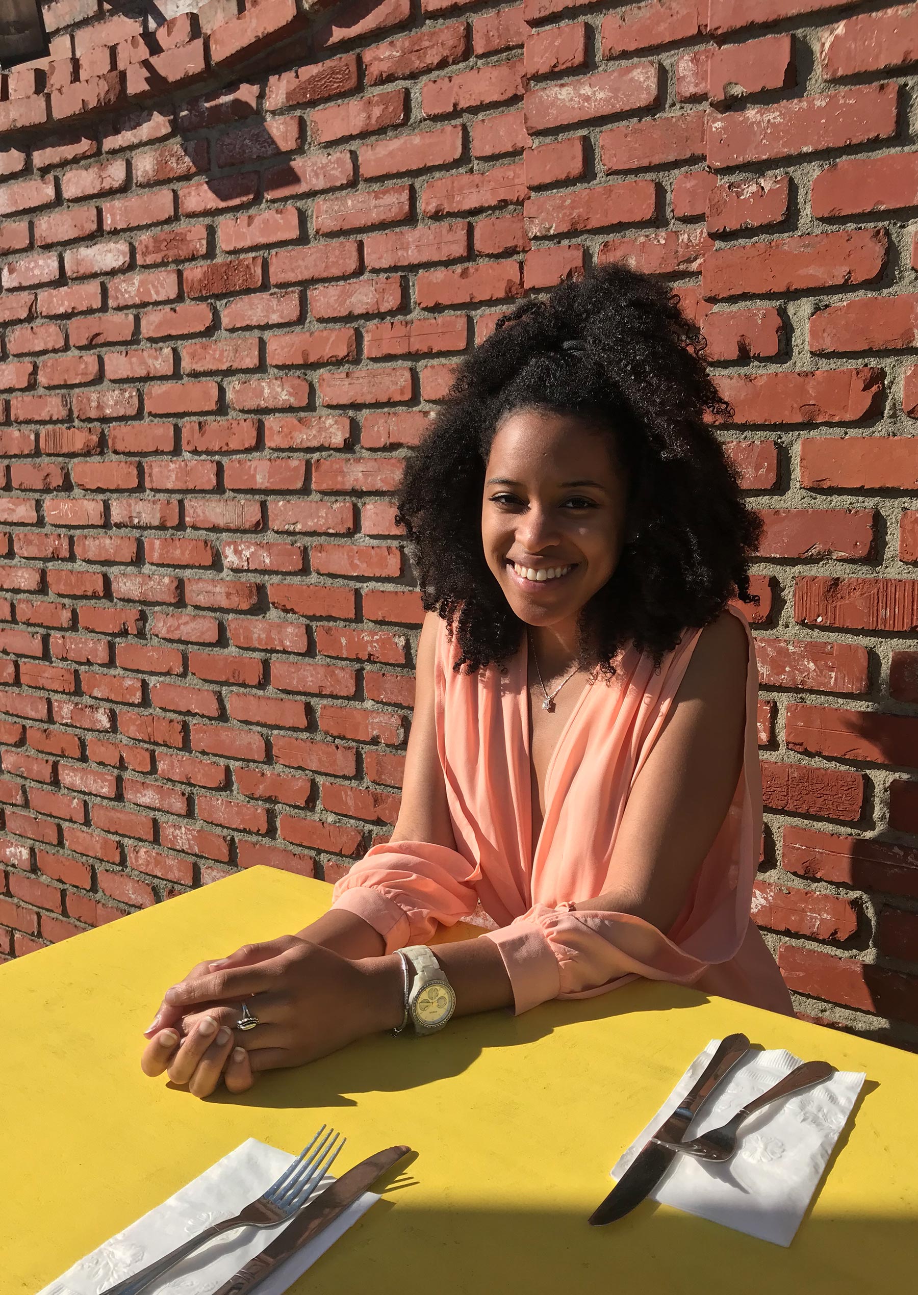 Lesley-Ann Agenor sits at a patio table in front of a bring wall, smiling