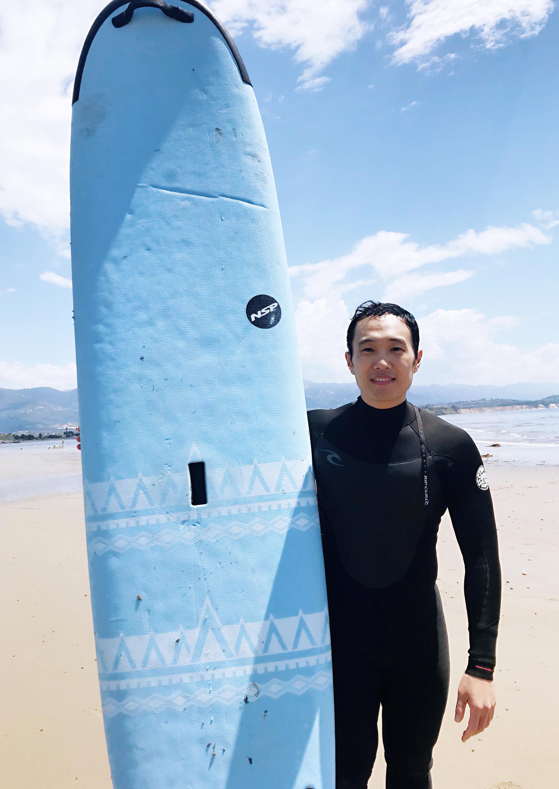 Michael Zhang wears a wetsuit and stands next to his surfboard on the beach