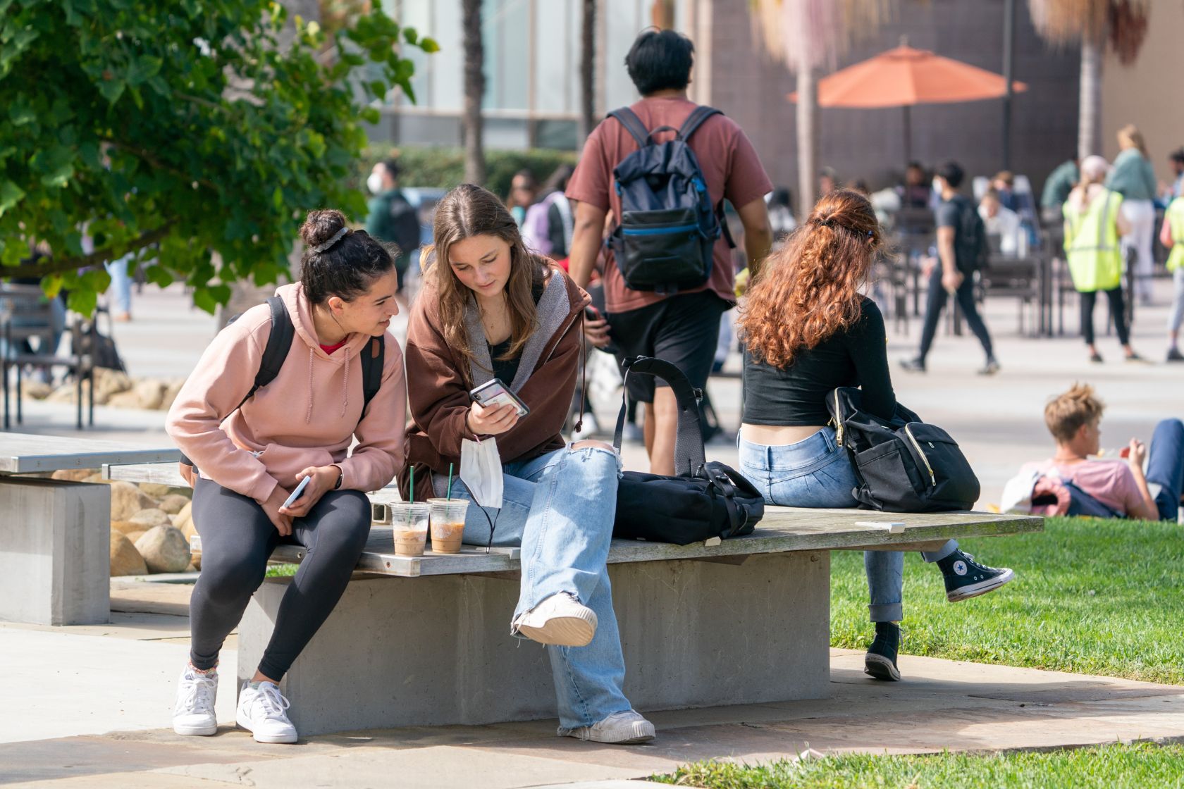 Two friends sit together with iced coffee drinks outside the UCSB Library with the busy Arbor behind them, looking down at one of their phones.