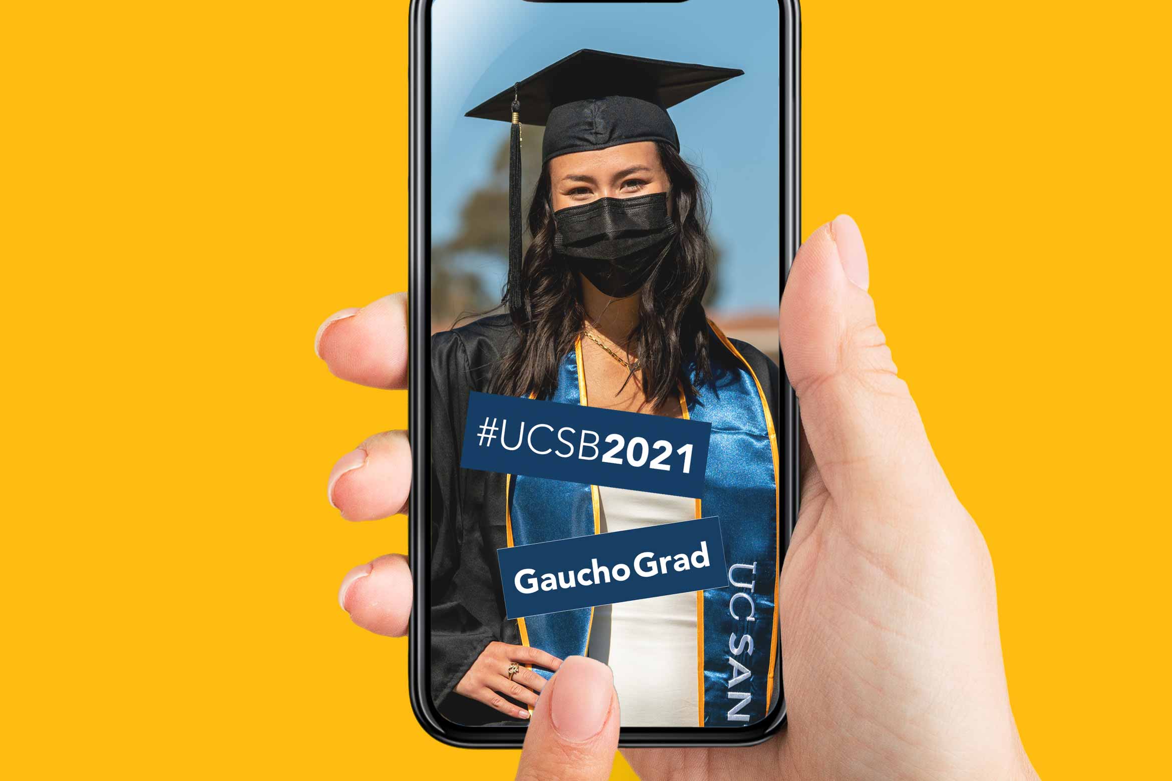 A photo of a hand holding a phone with a picture of a masked student wearing a cap and gown with GIPHY stickers placed on top that say "#UCSB2021" and "GauchoGrad"